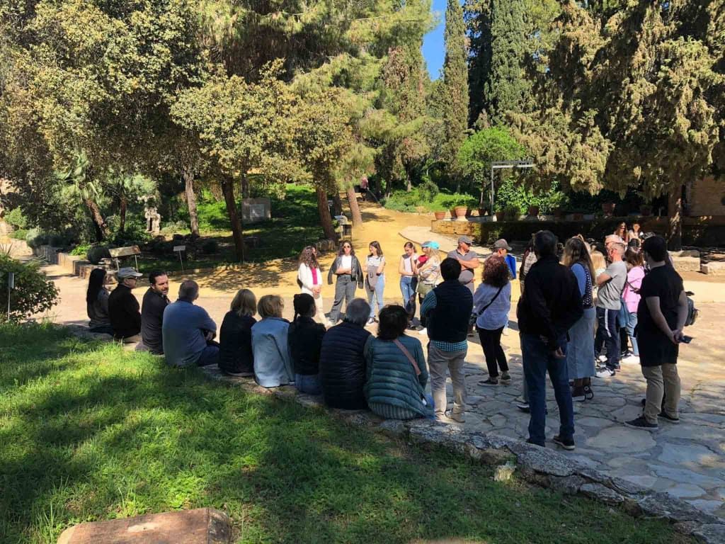Un grupo de personas en un parque escucha a un orador con los árboles al fondo.