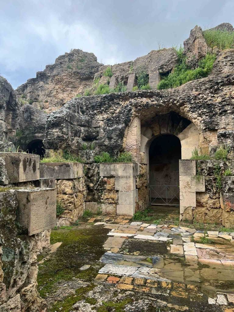 Ruinas de antiguas estructuras de piedra y arcos cubiertos de vegetación verde, bajo un cielo nublado.
