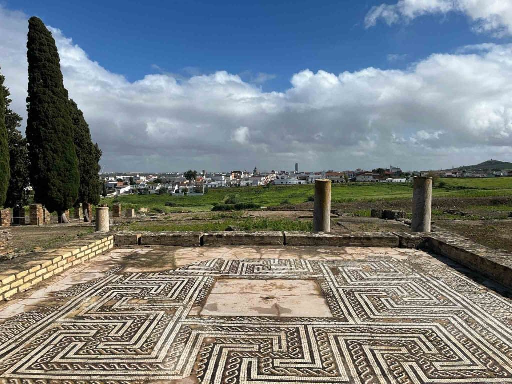 Suelo de mosaico en ruinas romanas con columnas, vista de un pueblo al fondo y un cielo azul con nubes.