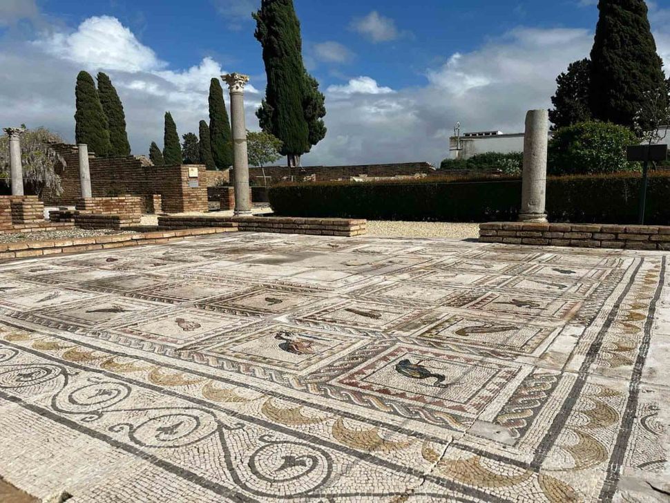 Suelo de mosaico romano antiguo con columnas, un seto y árboles bajo un cielo nublado en las ruinas, Sevilla.