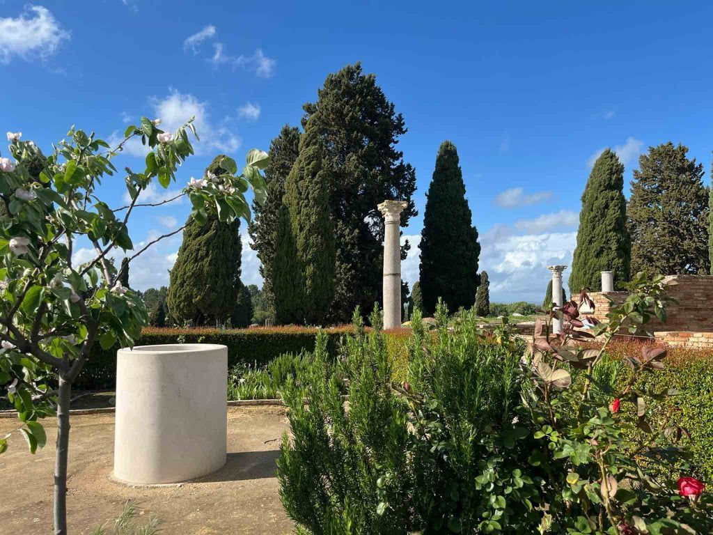 Un jardín soleado con altos cipreses, columnas de piedra y arbustos verdes bajo un cielo azul.