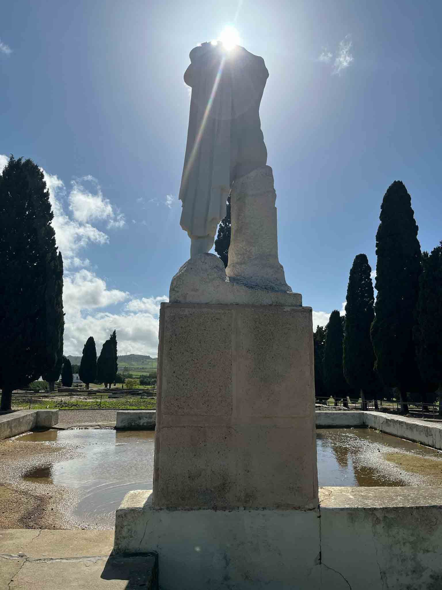 Estatua de una persona, recortada contra el sol, de pie sobre una base de piedra y árboles detrás de ella.