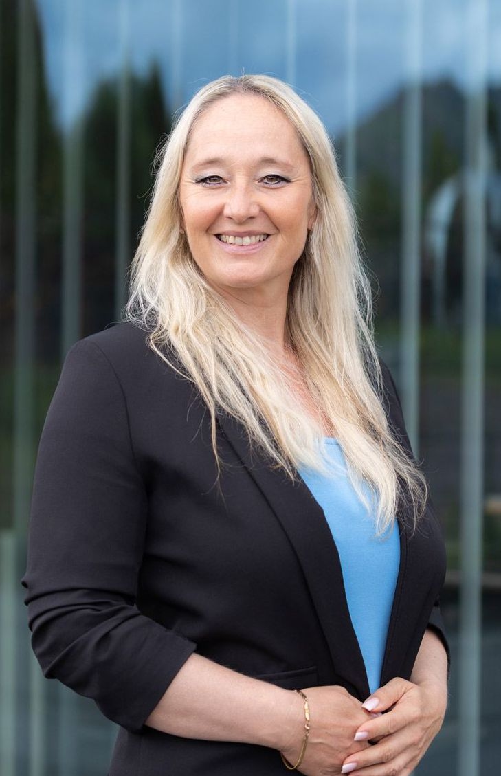 Blonde Frau im schwarzen Blazer lächelt vor einem Glasgebäude. Sie trägt ein blaues Oberteil und ein goldenes Armband.