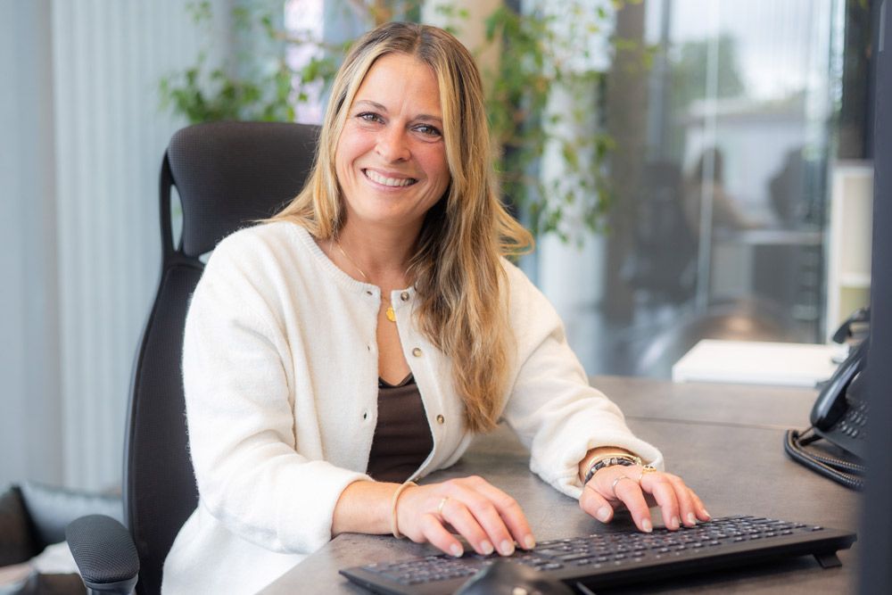 Frau mit langen blonden Haaren lächelt, während sie an einem Schreibtisch in einem modernen Büro tippt.