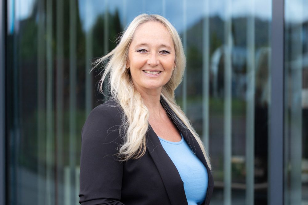 Blonde Frau im schwarzen Blazer lächelt in die Kamera, steht vor einem Glasgebäude.