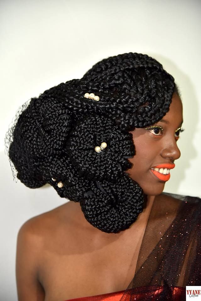 Femme avec une coiffure noire ornée de perles et vêtue d'un haut drapé marron, souriant de profil sur fond clair