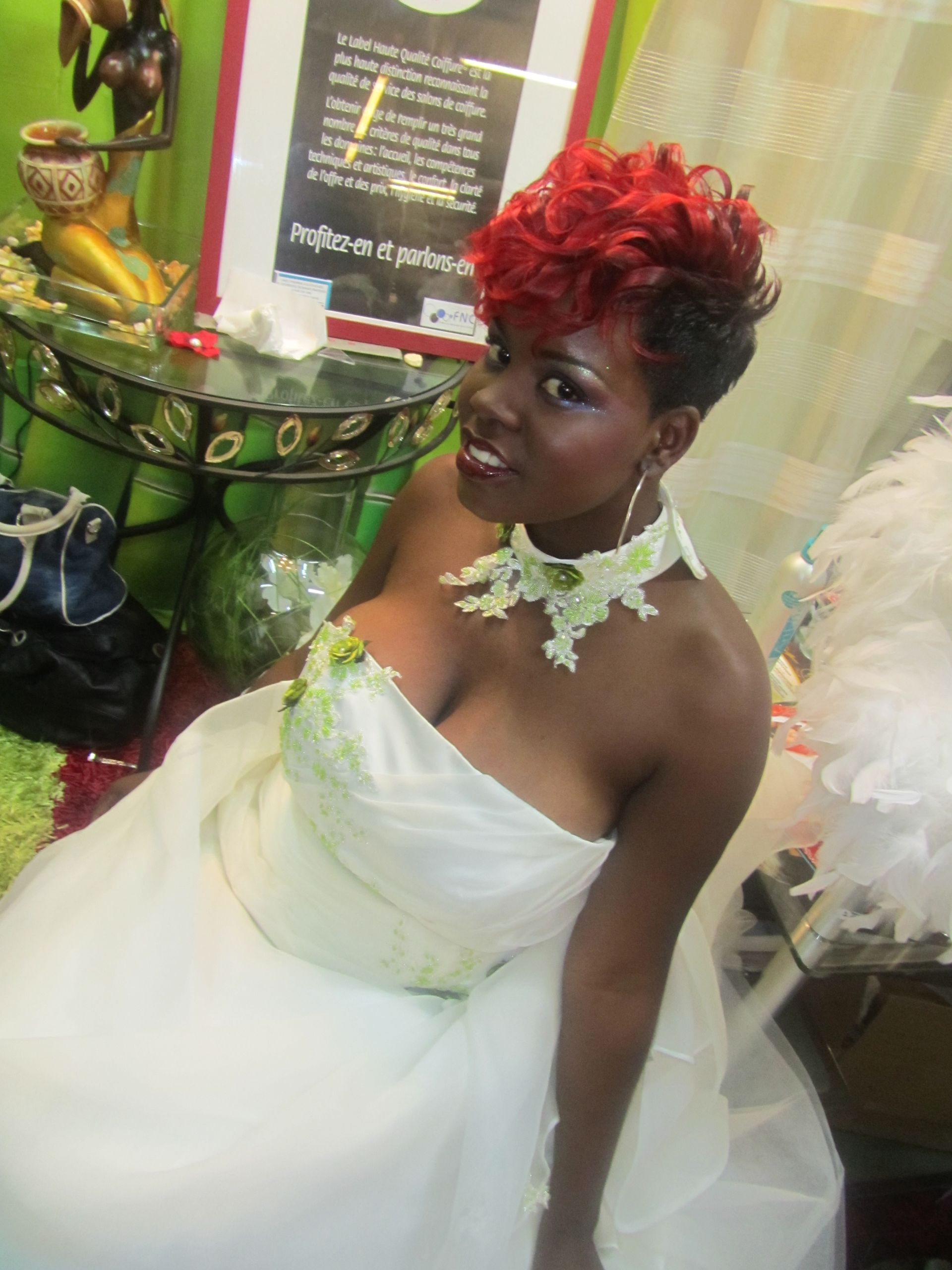 Une personne en robe blanche, aux cheveux rouges et portant un collier de fleurs, est assise à côté d'une table verte décorée.