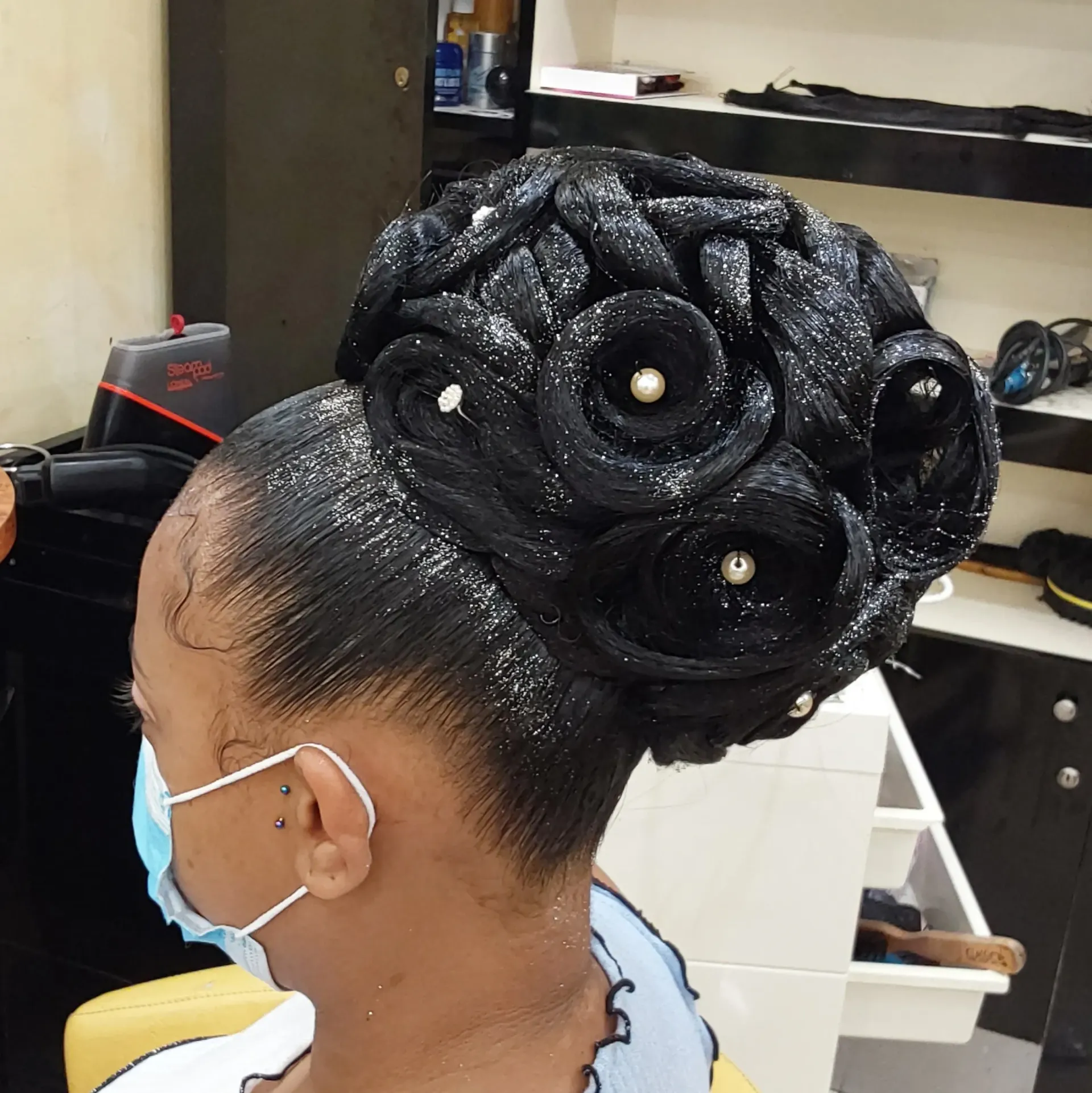 Coiffure réalisée en un chignon volumineux orné de roses noires pailletées et de perles, vue de profil dans un salon de coiffure.