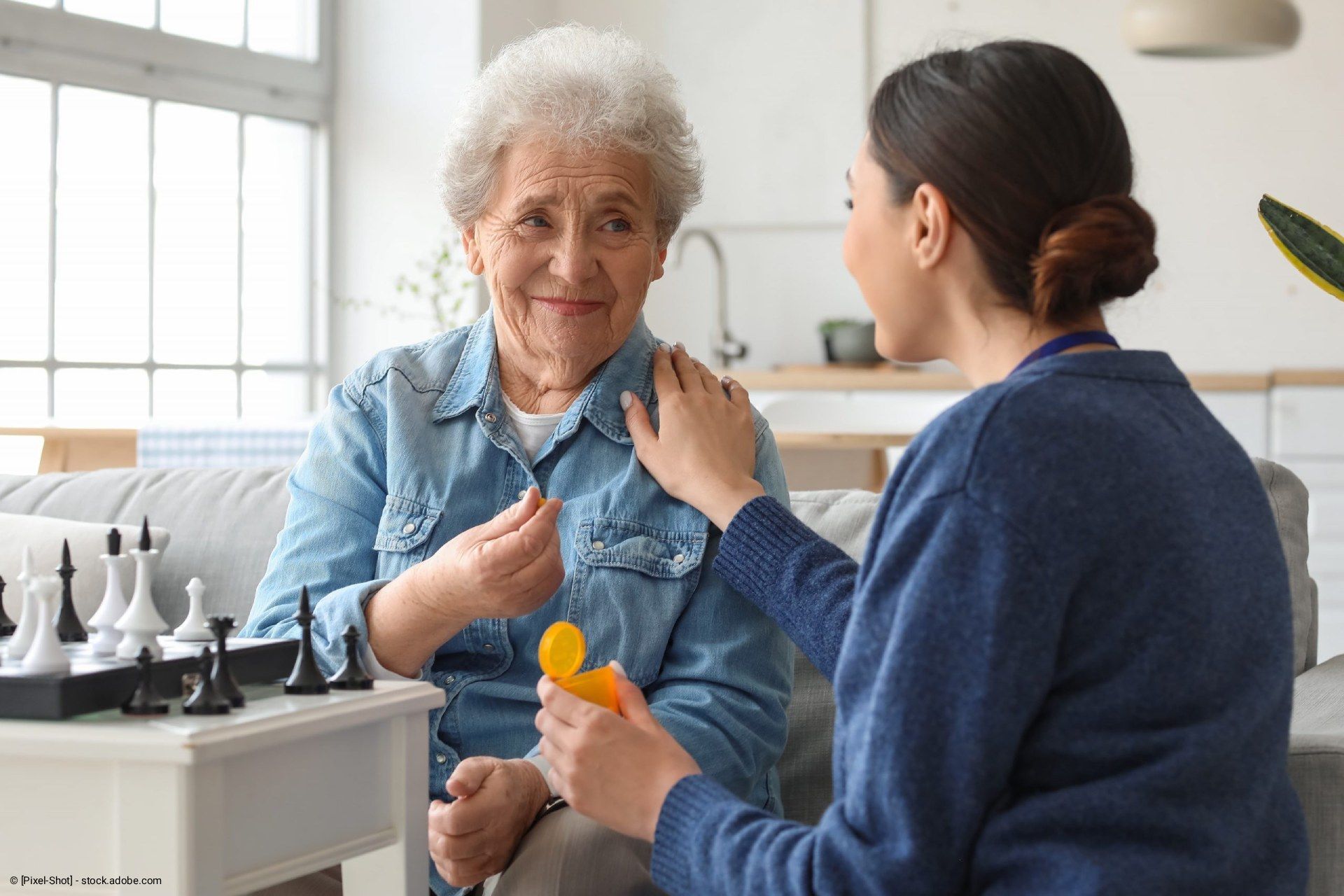 Eine Frau nimmt mit Unterstützung einer Pflegekraft Medikamente ein und lächelt. Ein Schachspiel liegt auf dem Tisch.