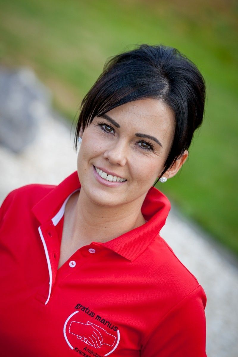 Frau in rotem Poloshirt lächelt im Freien. Logo auf dem Shirt; dunkles Haar, helle Haut.