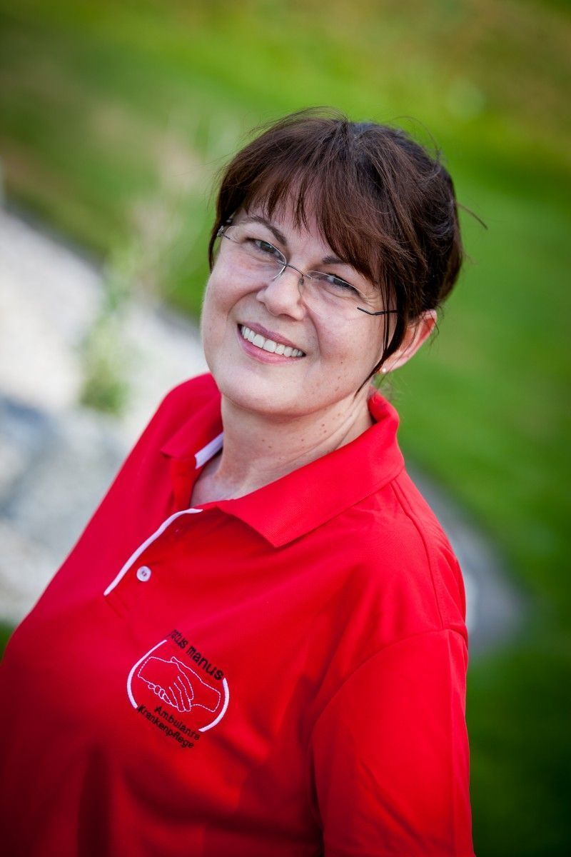 Frau lächelt im Freien; sie trägt eine Brille und ein rotes Poloshirt.