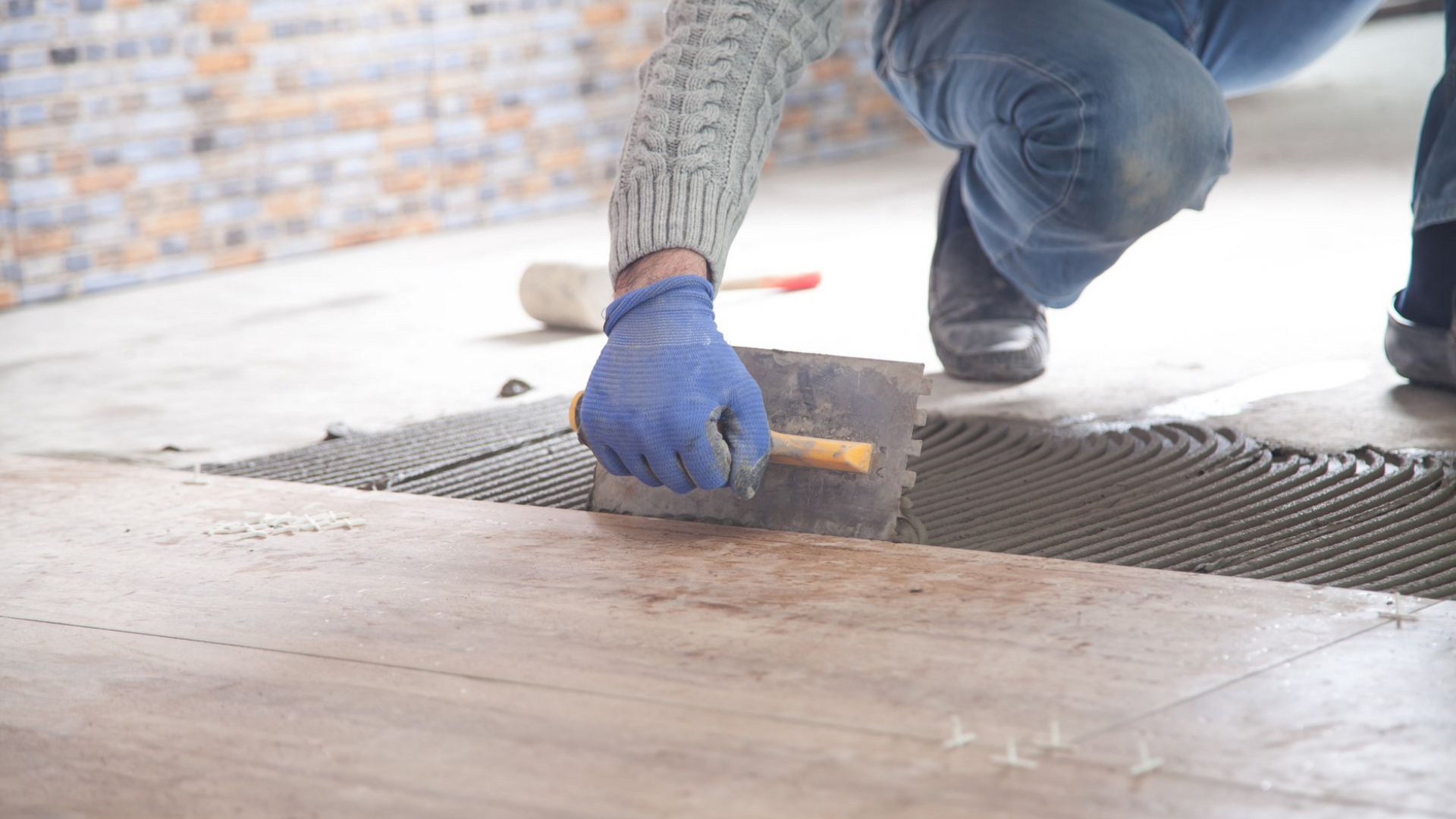 Personne agenouillée, appliquant du joint entre les carreaux de sol à l'aide d'une truelle, portant des gants bleus.