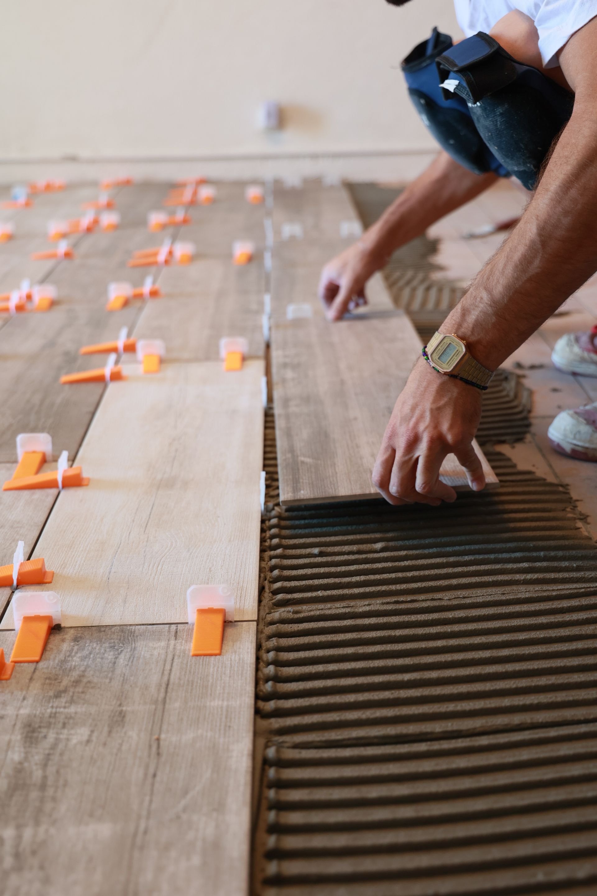 Un homme pose du carrelage imitation bois au sol à l'aide de clips de nivellement. On aperçoit les carreaux et les joints bruns.