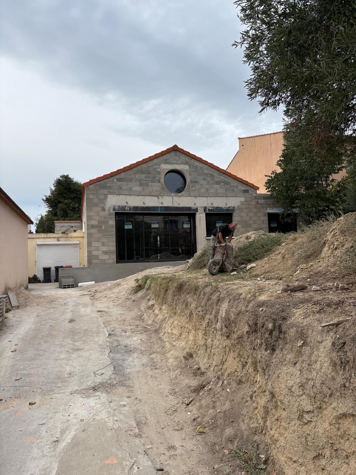 Chantier : bâtiment en béton inachevé avec de grandes fenêtres, une fenêtre ronde au-dessus. Une personne travaille à l'extérieur.