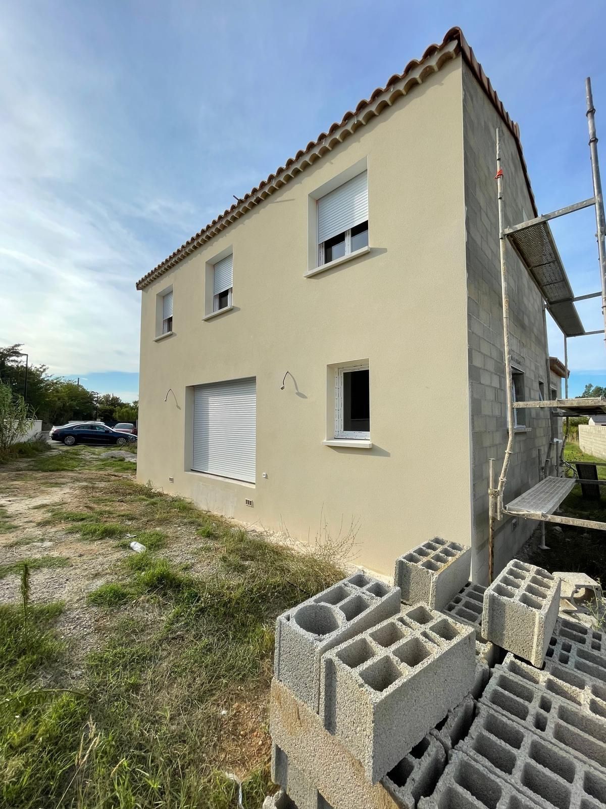 Maison de deux étages en construction, avec un enduit beige. Échafaudages, blocs de béton et ciel nuageux visibles.