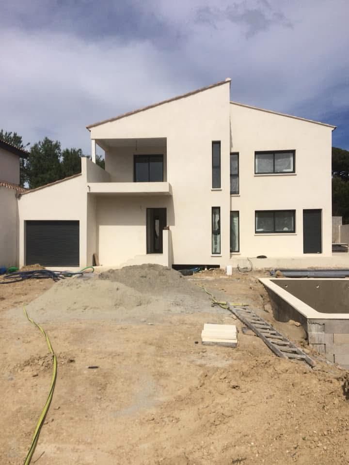 Maison blanche de deux étages en construction, avec garage, balcon et fenêtres à cadre sombre, se détachant sur un ciel nuageux.
