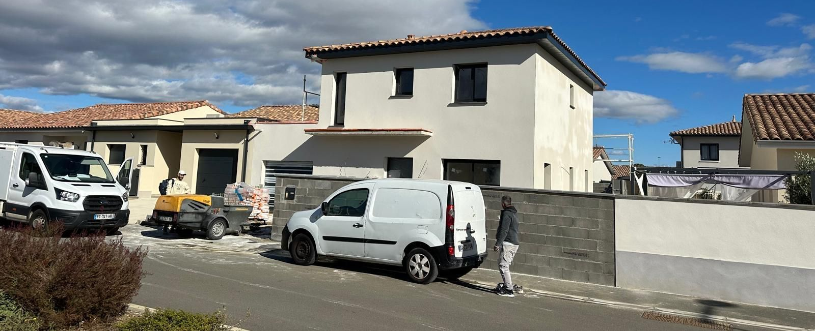 Chantier : camionnettes blanches, ouvriers, maison beige à deux étages, journée ensoleillée, ciel bleu, maisons aux toits de tuiles rouges.