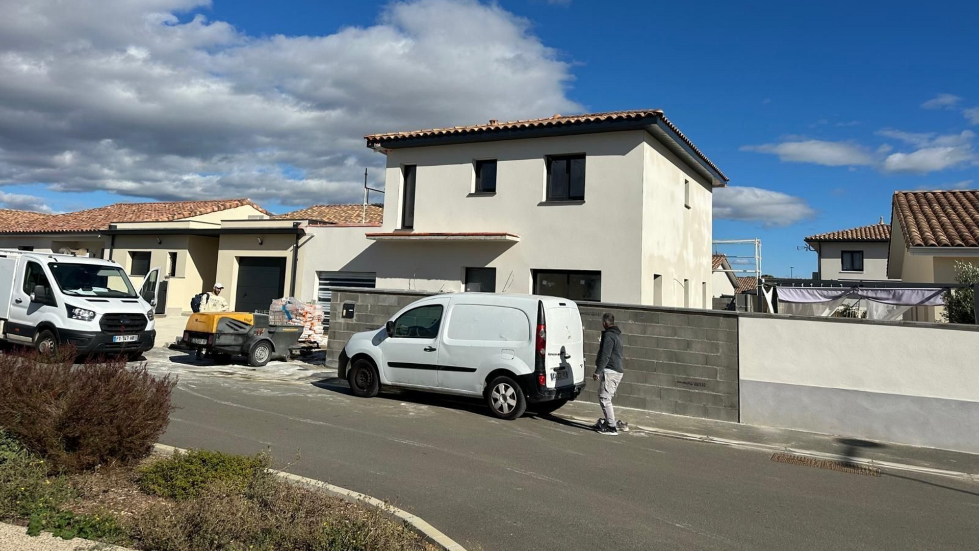 Chantier avec camionnettes et engins de chantier en bord de rue. Une maison à deux étages se dresse à l'arrière-plan. Journée ensoleillée.