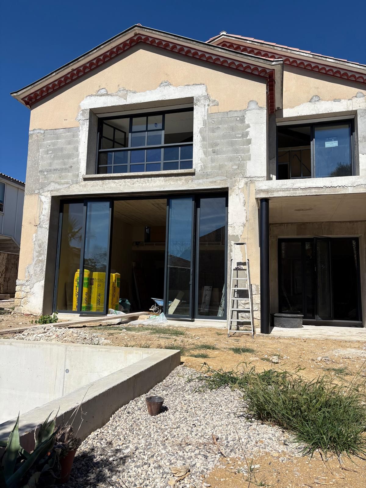Maison à deux étages en construction avec de grandes fenêtres et des portes coulissantes, cadre extérieur ensoleillé.