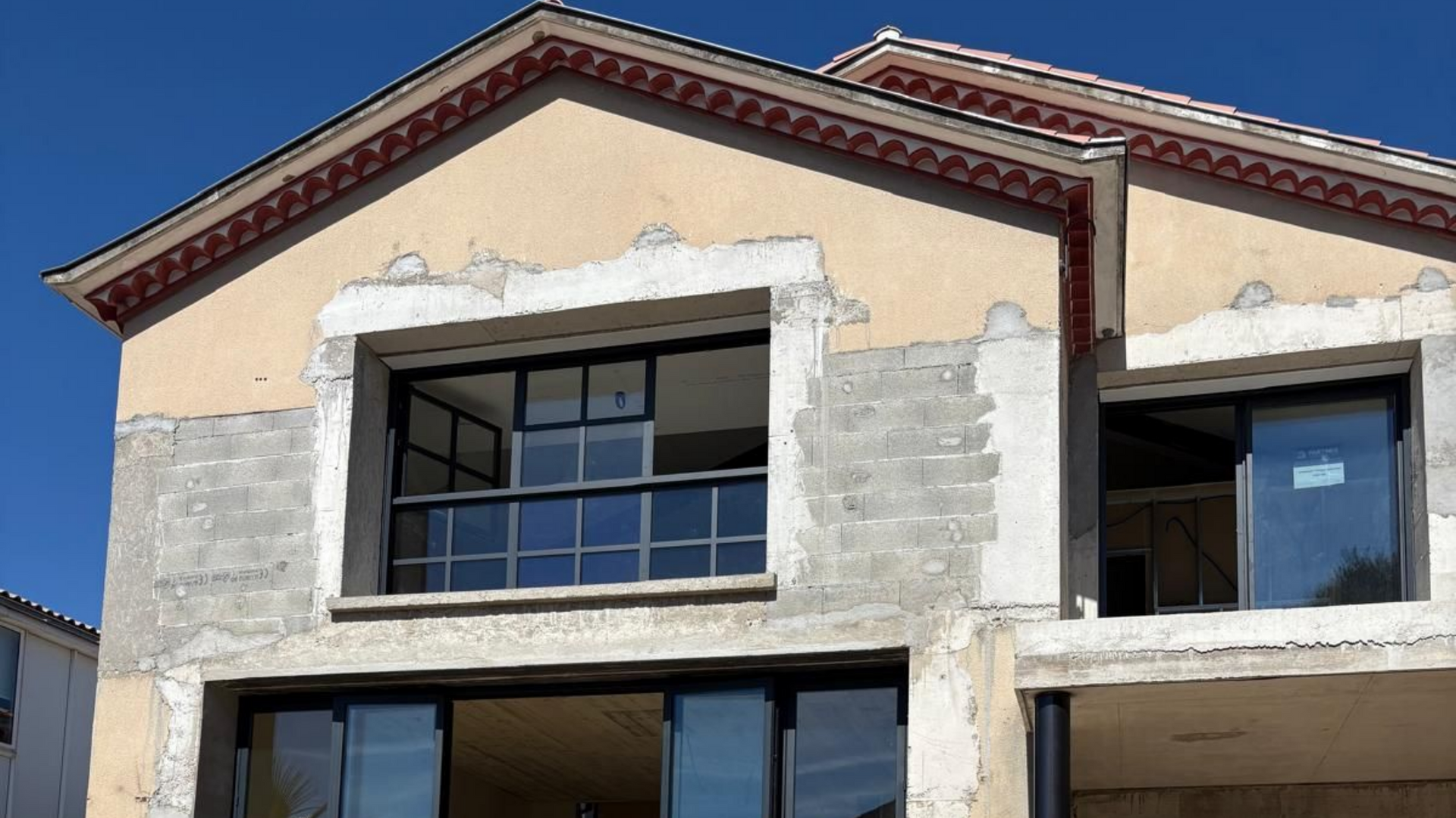 Extérieur d'un bâtiment en construction, avec des fenêtres à cadre noir et une façade en stuc inachevée.