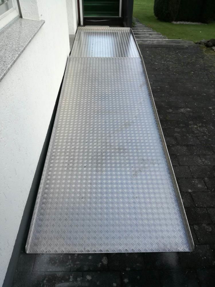 Eine Metallrampe, die zu einer Tür führt, im Hintergrund eine Treppe