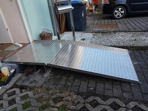 Auf einem gepflasterten Gehweg steht neben einem Auto eine Metallrampe.