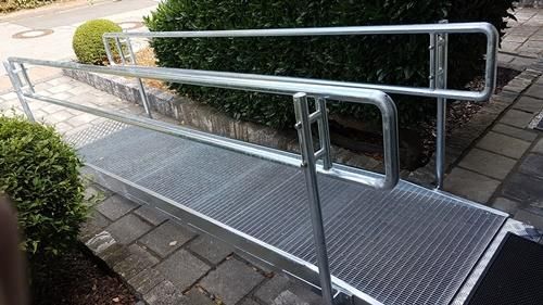 Auf einem Gehweg neben einem Busch steht eine Metallrampe mit Geländer.