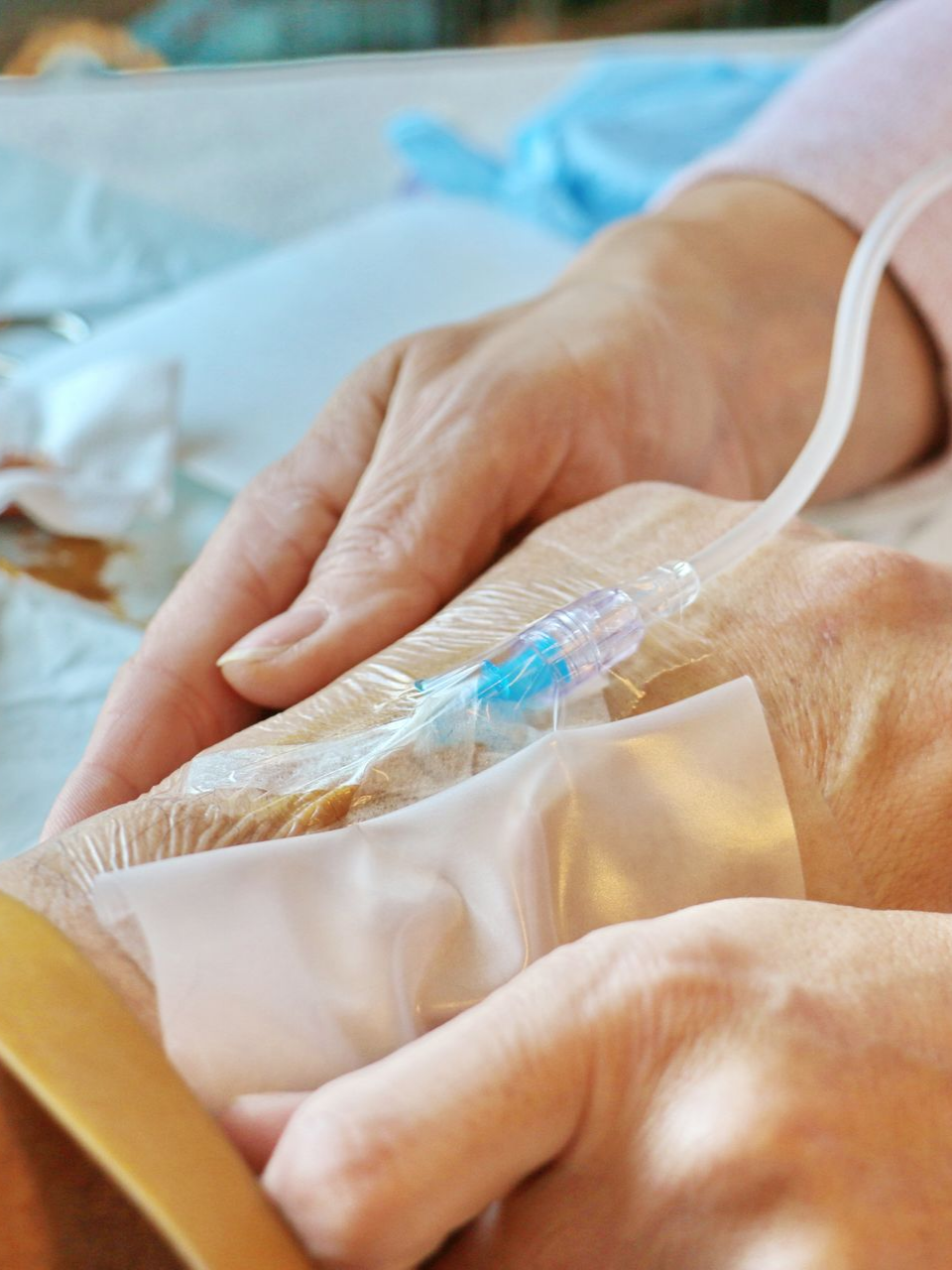 Deux mains tiennent la main d'un patient perfusé. On aperçoit du ruban adhésif blanc et une tubulure transparente.
