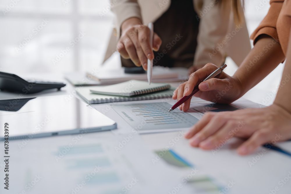 Zwei Personen sitzen an einem Tisch und sichten Dokumente. Hände deuten auf Diagramme und Grafiken; Notizbücher und ein Tablet liegen daneben.