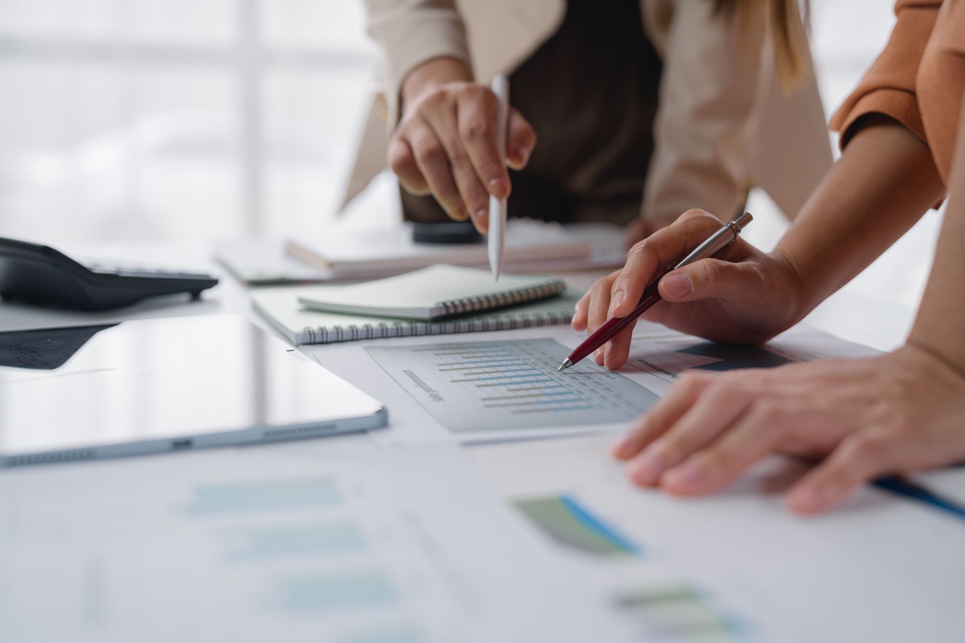 Zwei Personen sitzen an einem Tisch und sichten Dokumente. Hände deuten auf Diagramme und Grafiken; Notizbücher und ein Tablet liegen daneben.