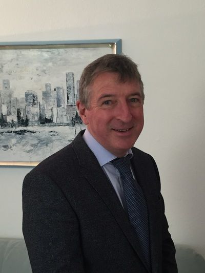 Mann im Anzug, lächelnd, vor einem Stadtbild-Gemälde.