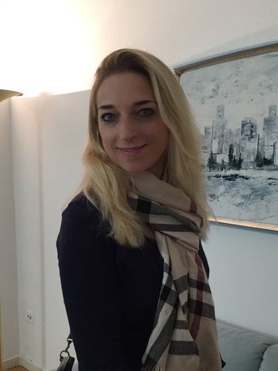 Blonde Frau lächelt, trägt einen Schal und ein dunkles Oberteil; sie steht in einem Innenraum in der Nähe eines Stadtbildgemäldes.