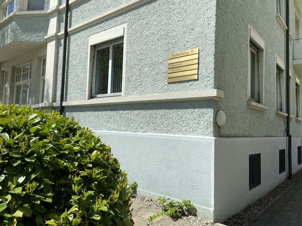 Hellblaues Gebäude mit Fenster und goldener Plakette an der strukturierten Fassade, Busch davor.
