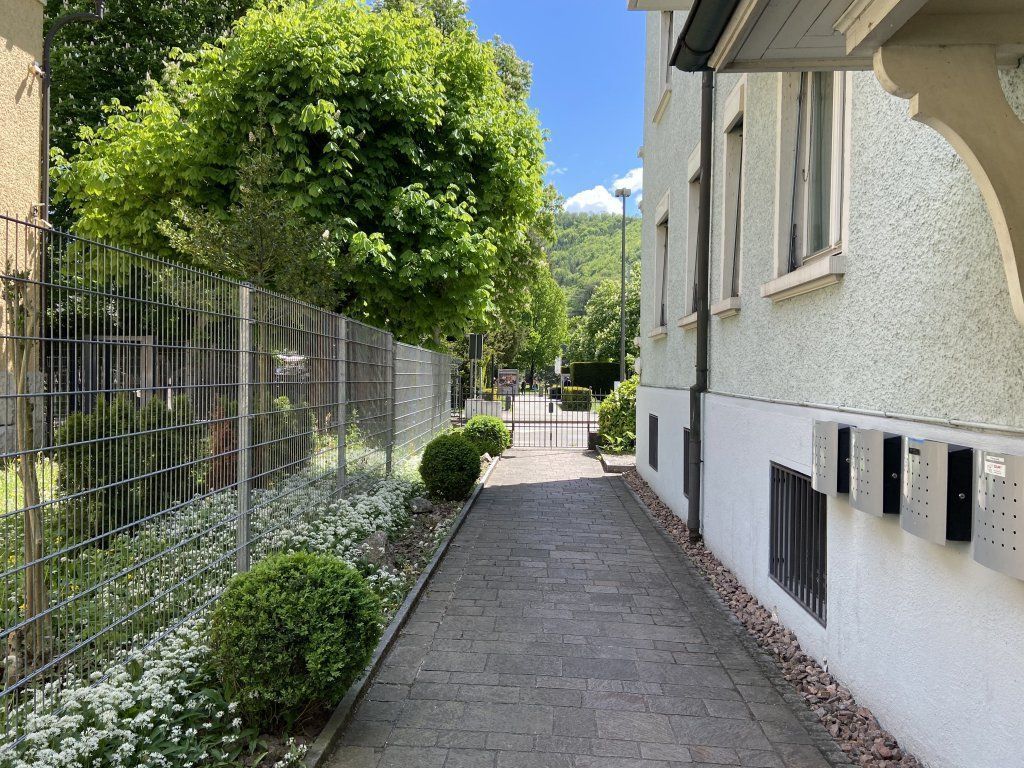 Ein schmaler Gehweg zwischen einem Gebäude und einem Zaun, der zu einer Strasse führt, an einem sonnigen Tag.