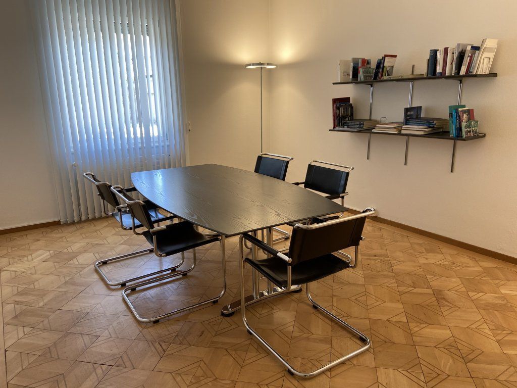 Konferenzraum mit Tisch, Stühlen, Fenster, Stehlampe und Wandregalen mit Büchern.