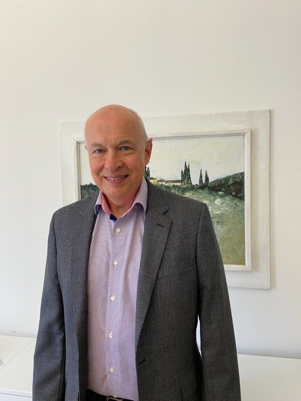 Ein Mann in grauem Blazer und offenem Hemd lächelt vor einem Gemälde.