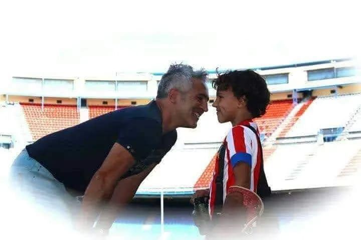Un adulto y un niño con camiseta de fútbol se enfrentan en un estadio vacío, sonriendo y compartiendo un momento cercano.