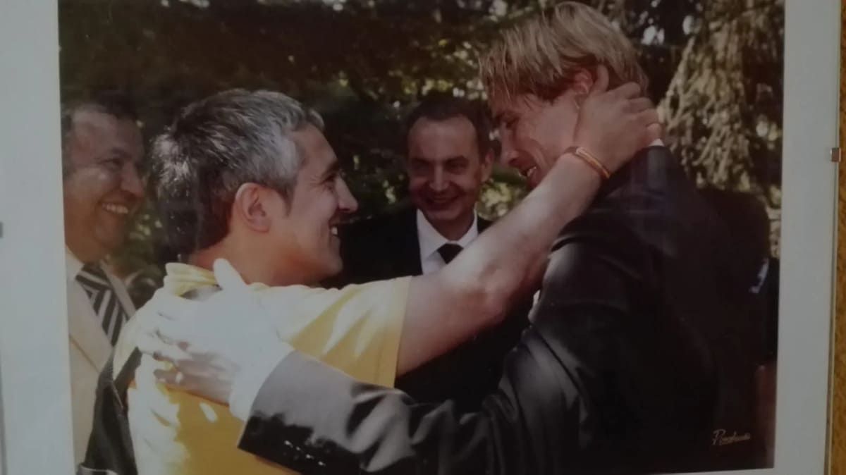 Una persona con una camisa amarilla y guantes blancos abraza a una persona con un traje oscuro, mientras otra persona observa al fondo.