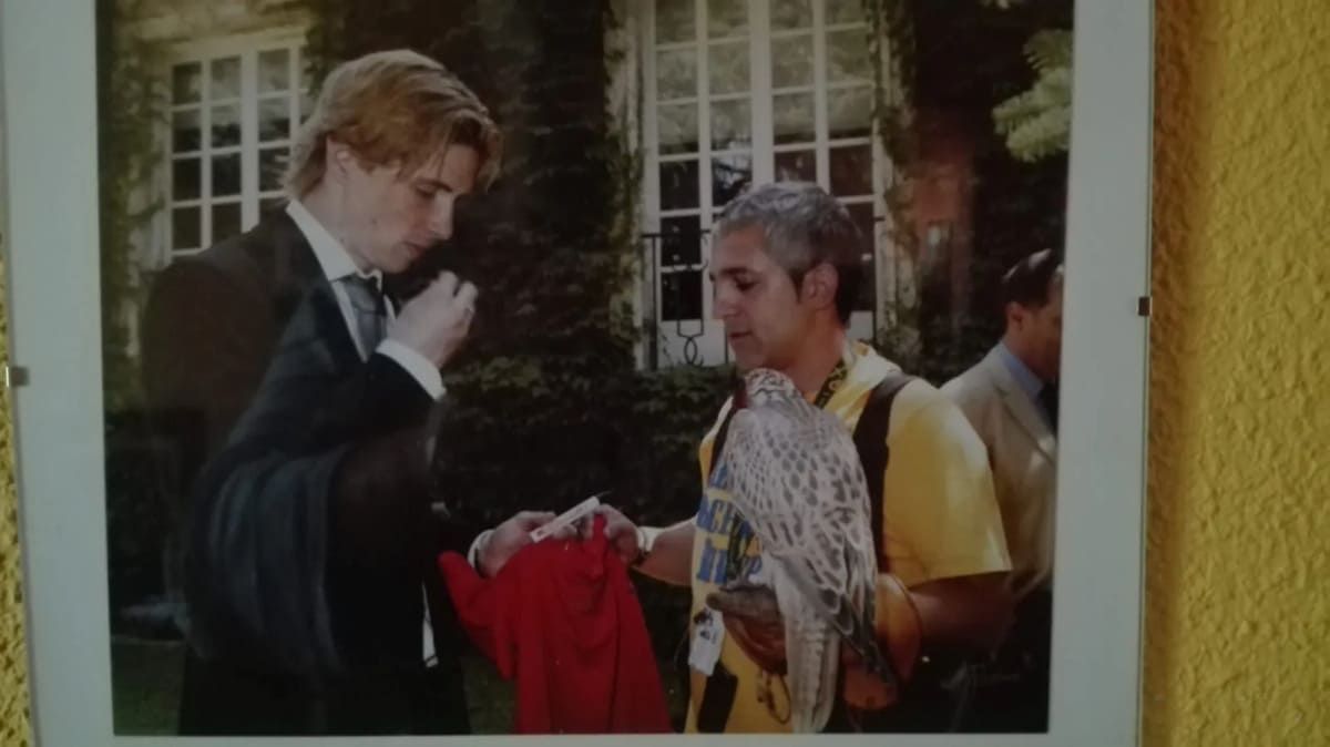 Una persona rubia con traje y una persona con camisa amarilla sostienen un ave rapaz en un jardín.