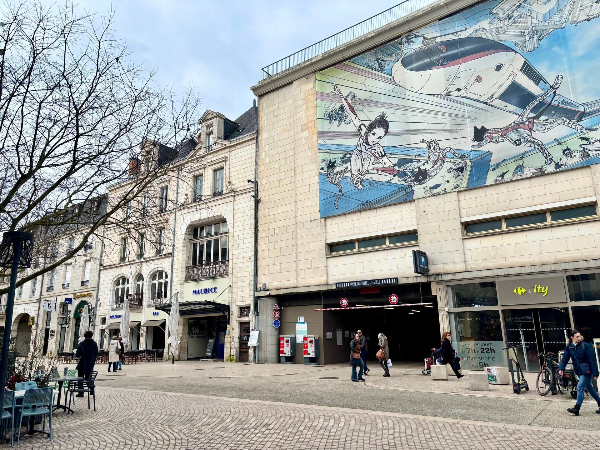 Une place pavée devant des bâtiments historiques et une structure moderne ornée d'une grande fresque murale.