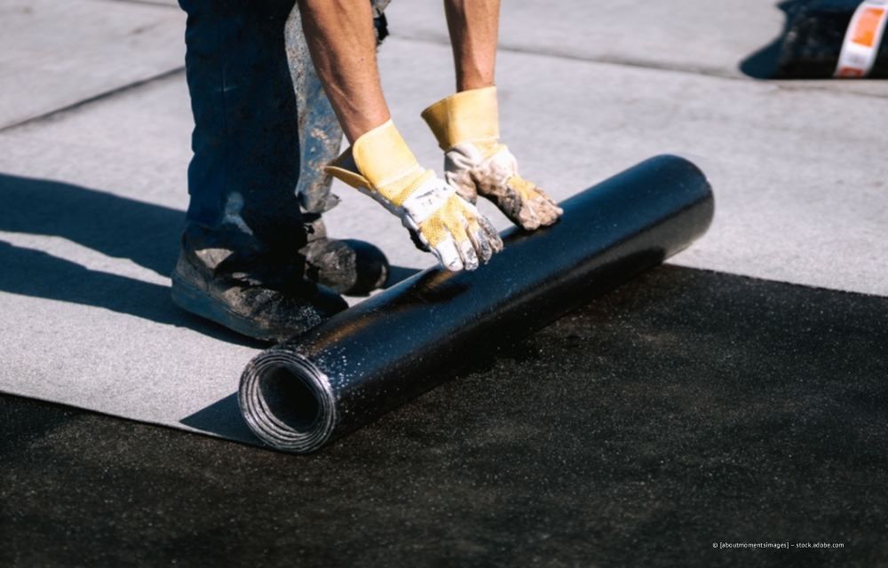 Ein Arbeiter rollt mit behandschuhten Händen schwarzes Dachmaterial auf einem Flachdach aus.