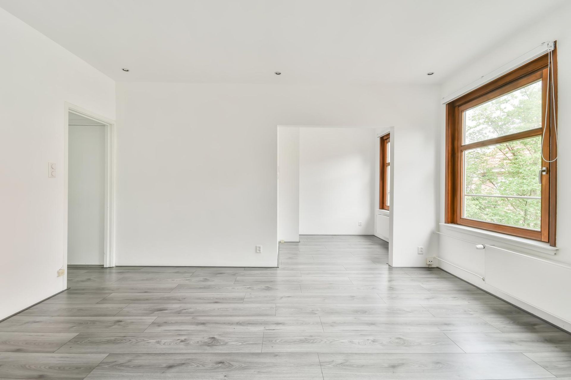 Habitación blanca vacía con suelo de madera gris y grandes ventanales que dejan entrar luz natural.