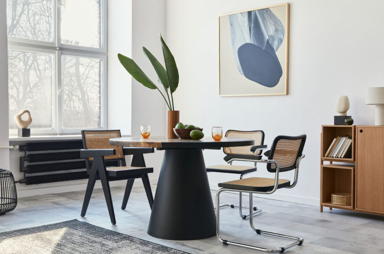 Comedor moderno con mesa negra, sillas de mimbre, ventana grande, planta y arte abstracto en la pared.