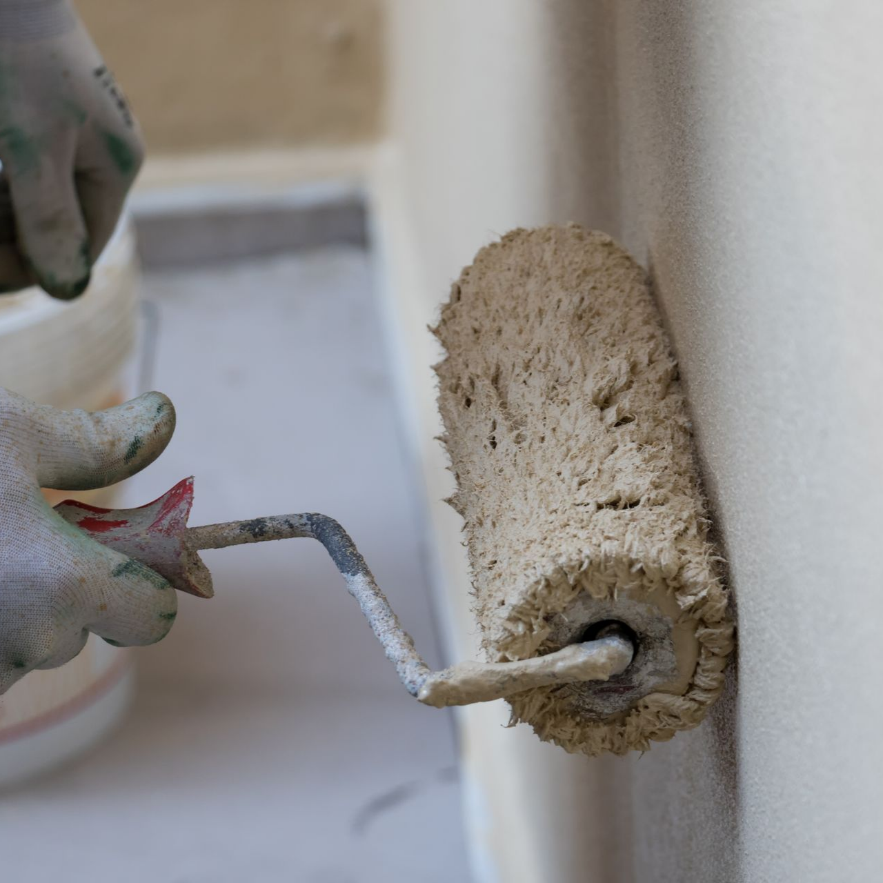 Une personne peint un mur beige avec un rouleau à peinture, portant des gants.