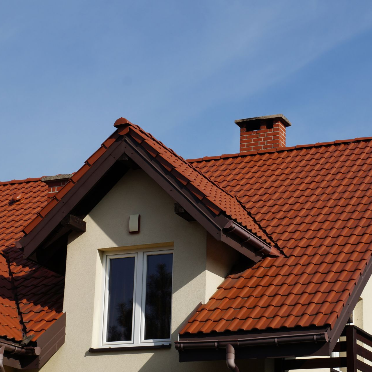 Toit de tuiles rouges sur une maison de couleur crème avec une cheminée contre un ciel bleu.