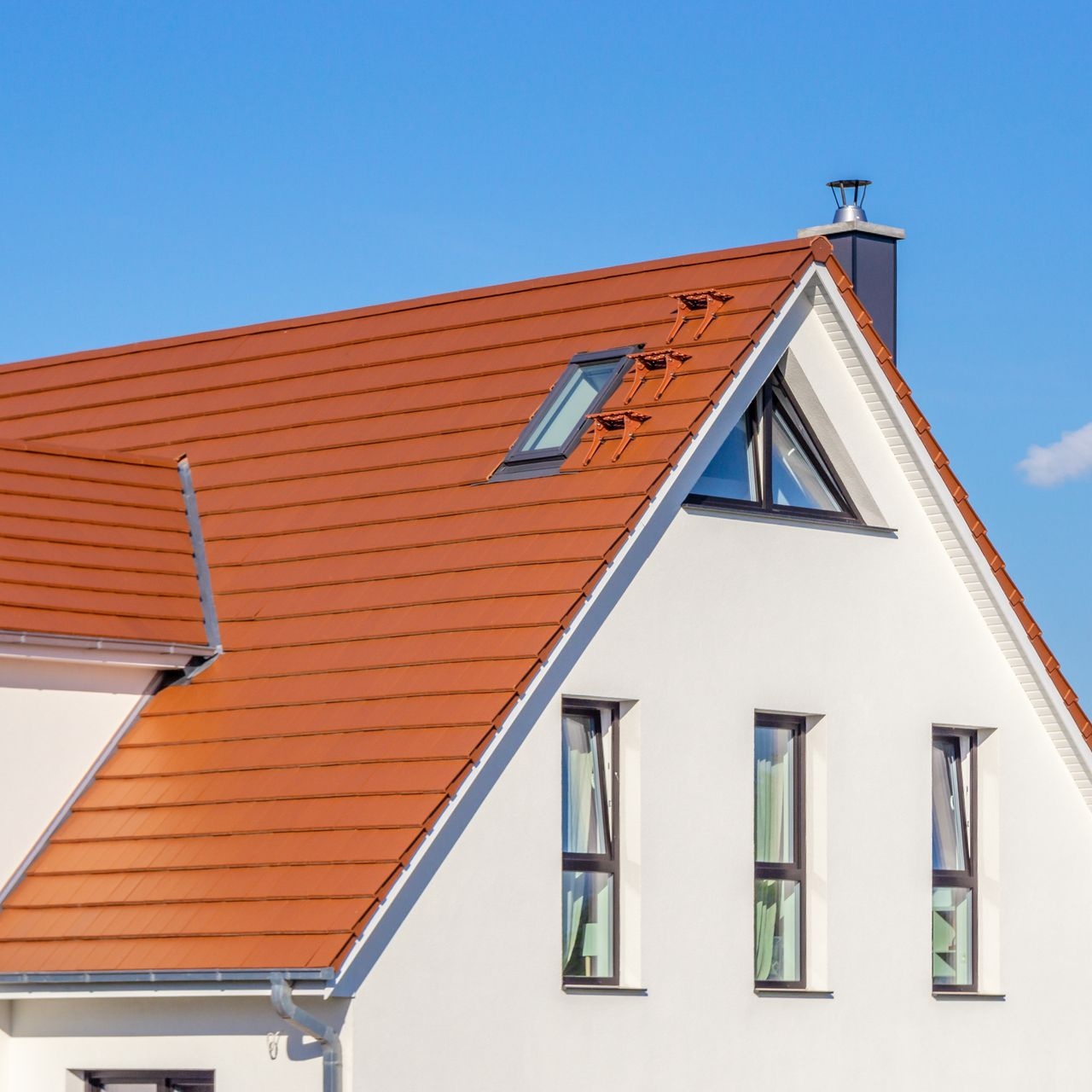 Maison blanche avec un toit de tuiles orange, trois fenêtres, une lucarne et une cheminée contre un ciel bleu.