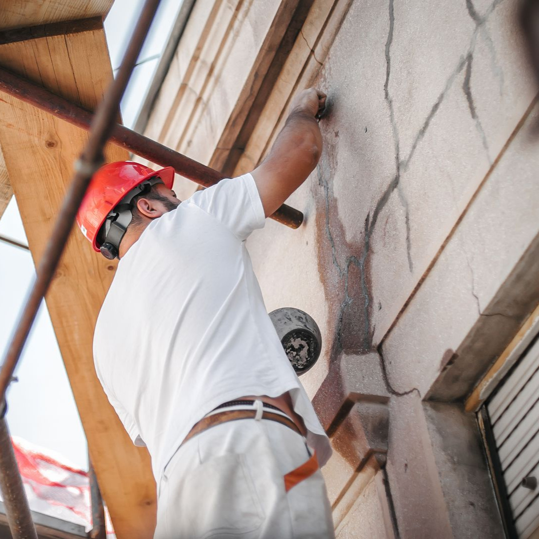 Un homme portant un casque répare un mur en stuc, debout sur un échafaudage.