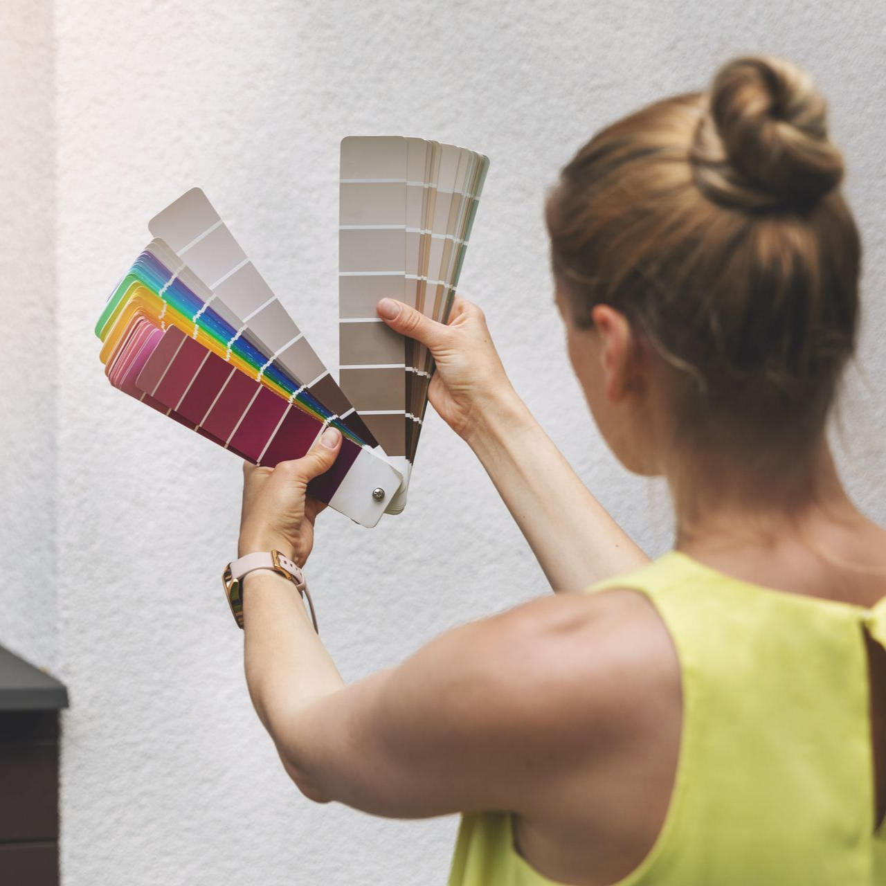 Une femme tenant des échantillons de couleurs de peinture, comparant les couleurs à un mur blanc à l'extérieur.