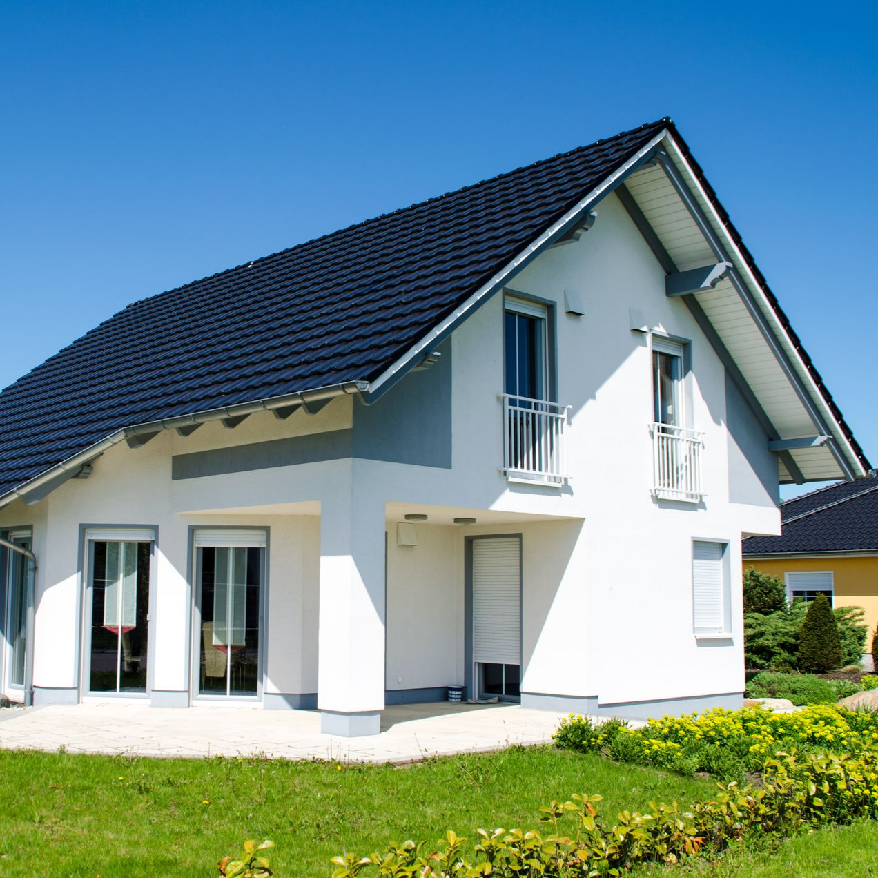 Maison blanche avec toit noir, petits balcons et pelouse verte par une journée ensoleillée.