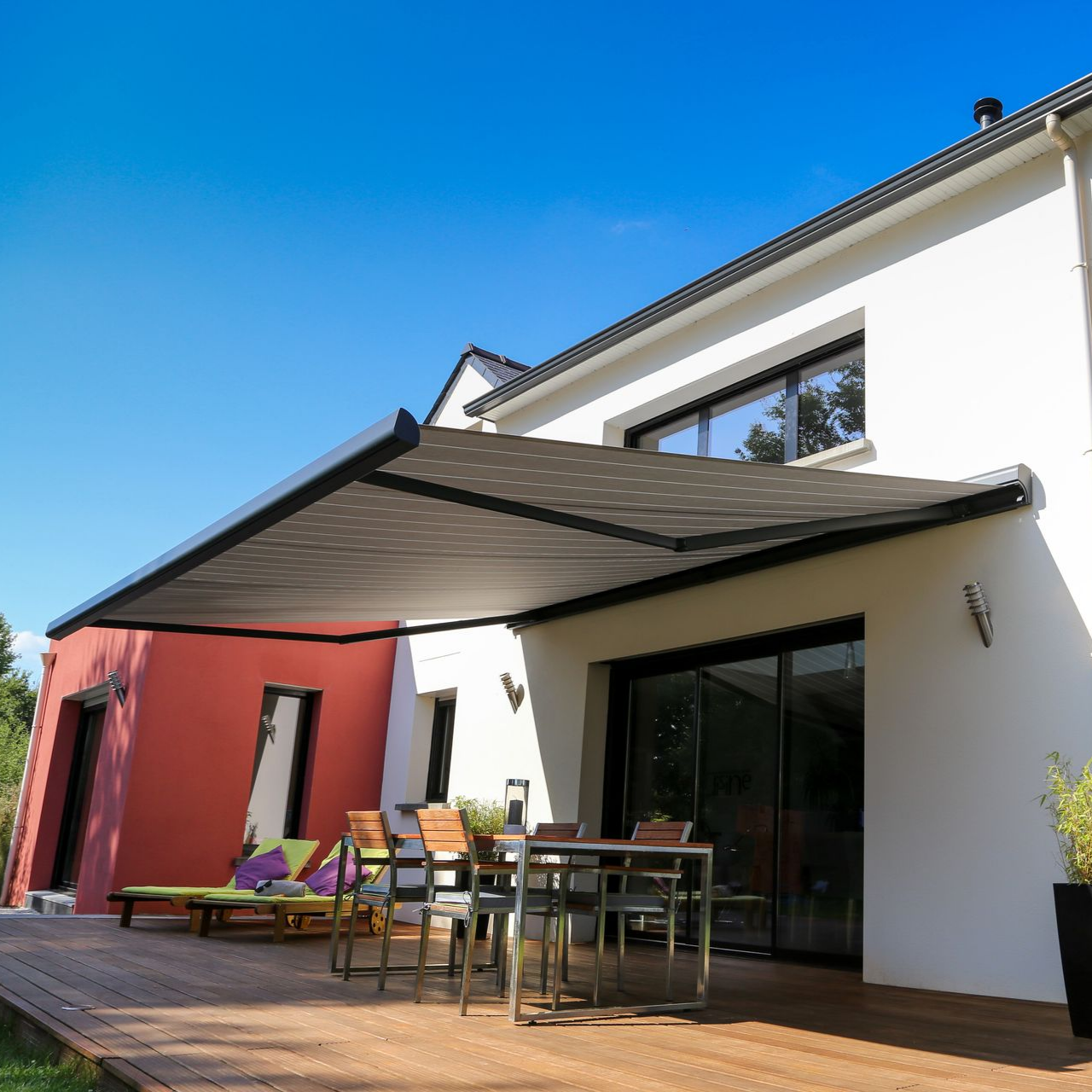 Terrasse extérieure avec auvent rétractable au-dessus d'une table à manger, de chaises longues et d'une terrasse en bois à côté d'une maison blanche avec de grandes fenêtres.